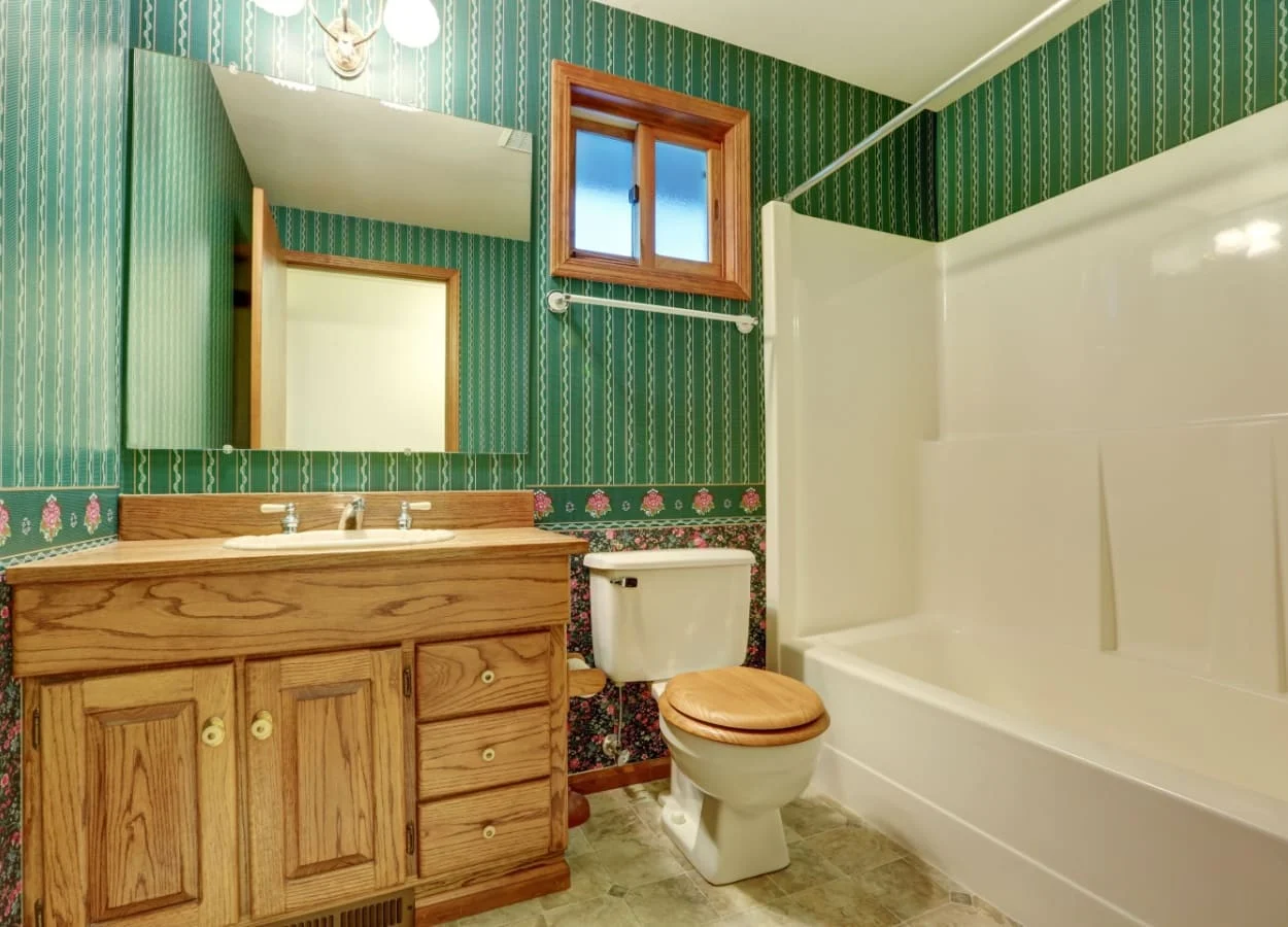  baie in stil vintage, amenajata cu mobilier din lemn, tapet decorativ verde cu model floral si cada cu panou din acril 